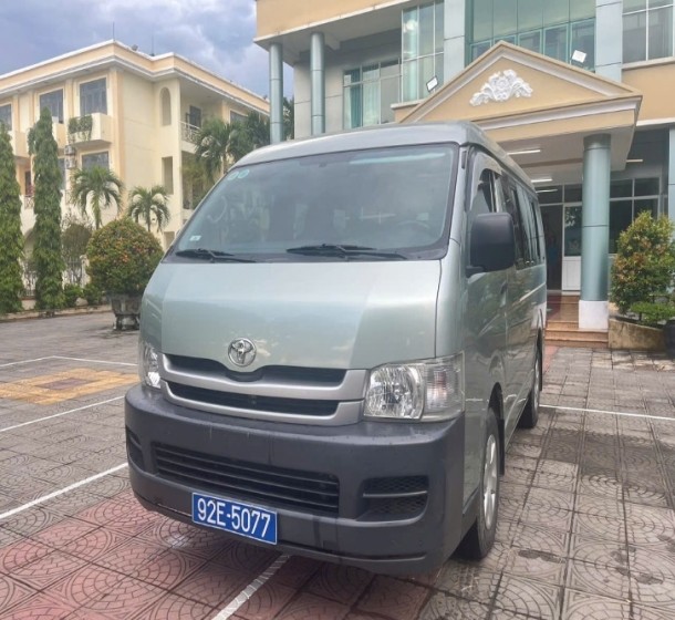 Khoang lái của xe khách 16 chỗ Toyota Hiace đời cũ, nhìn từ ghế hành khách, hiển thị bảng điều khiển và vô lăng.