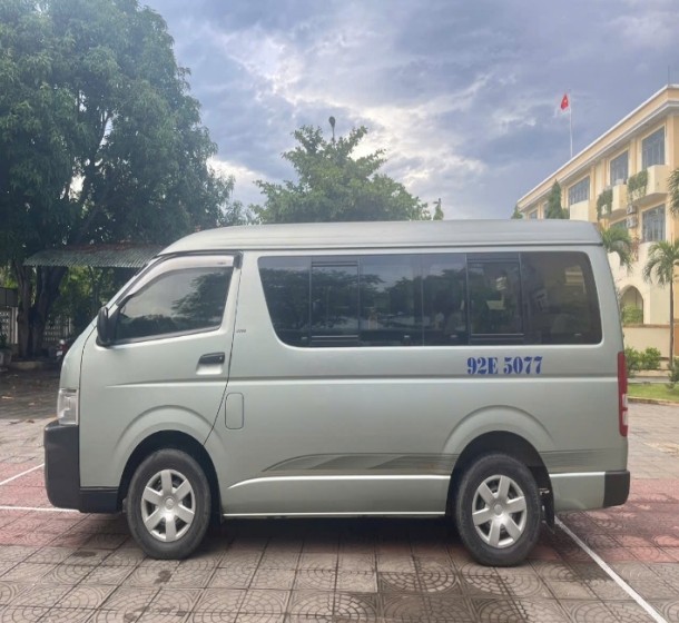Hình ảnh một chiếc xe ô tô khách cũ 16 chỗ Toyota Hiace màu trắng, biển số 92E-5077, bên ngoài thân xe có vài vết trầy xước nhẹ.