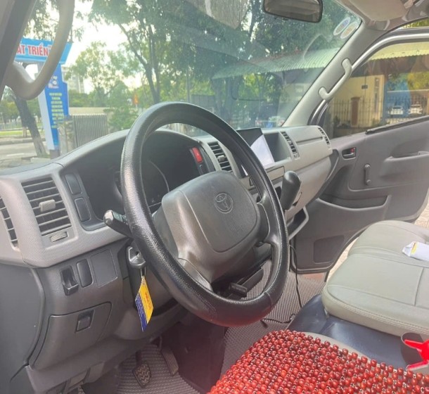 Thân xe Toyota Hiace 16 chỗ cũ màu trắng với cửa trượt bên hông mở ra, lộ nội thất ghế ngồi, cho thấy độ rộng rãi.