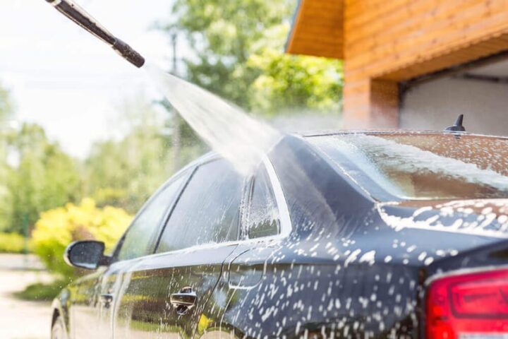 Người đang tự rửa xe ô tô tại nhà với vòi xịt nước áp lực cao, chuẩn bị cho quy trình vệ sinh chuyên nghiệp